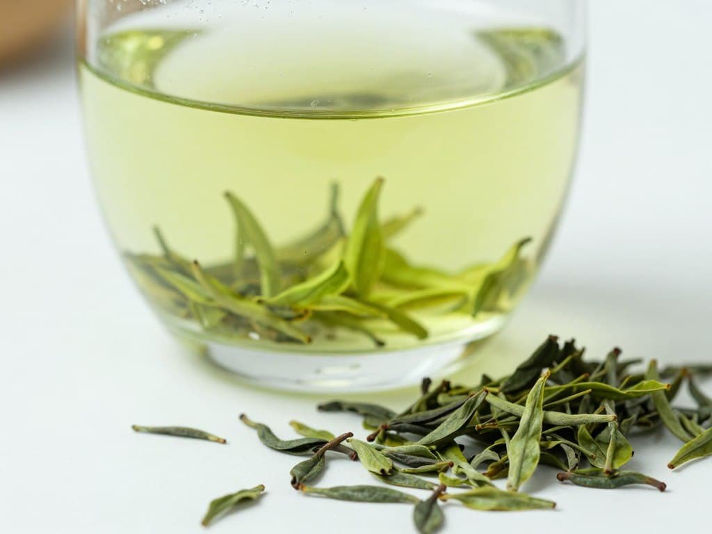 Fresh green tea in a clear glass cup with loose green tea leaves scattered around on white background Fresh green tea in a clear glass cup with loose green tea leaves scattered around on white background
